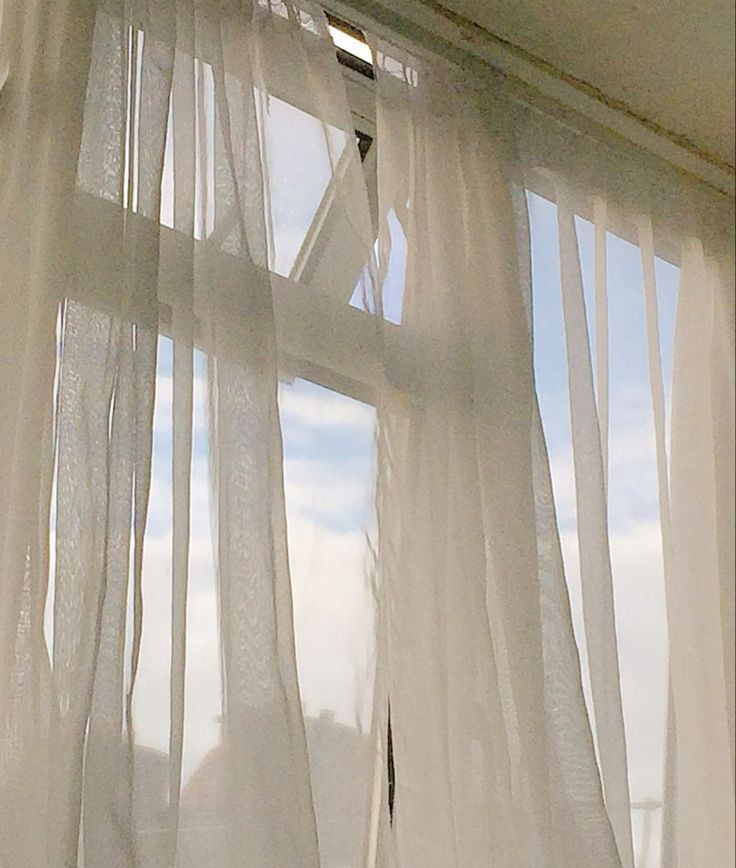 A window with sheer white curtains softly blowing, revealing a glimpse of blue sky and clouds.